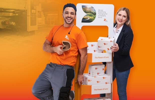 dunkelhaariger junger Mann steht links neben einem Aufsteller aus Wellpappe, freundlich in die Kamera lächelnd, mit einem kleinen Wellpappkarton in der Hand. Mütze und Gehörschutz und oranges T-Shirt, Arbeitskleidung, vor orangefarbenem Hintergrund. Rechts vom Aufsteller steht eine junge blonde Frau mit langen Haaren und dunklem Jacket und lächelt in die Kamera