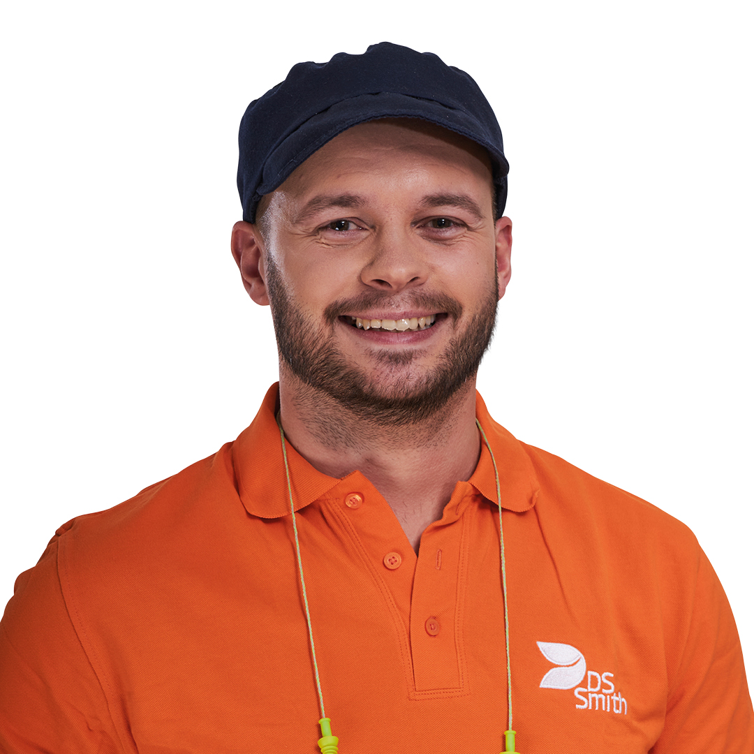 Portrait eines jungen Mannes, Bart, Mütze und Gehörschutz mit orangenem T-Shirt, Arbeitskleidung der Produktion. Er lächelt in die Kamera.