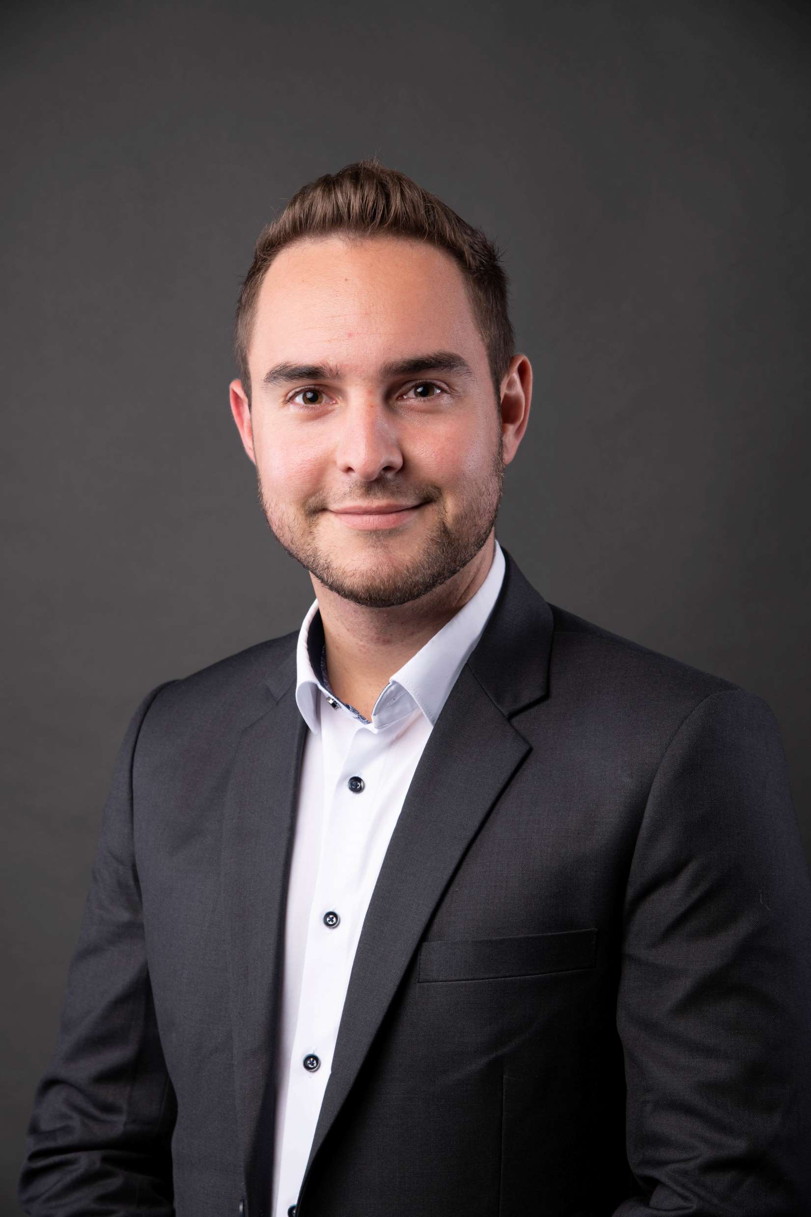 Portrait eines jungen Mannes mit kurzen, dunklen Haare. Weißes Hemd, dunkles Jacket. Er schaut in die Kamera. 