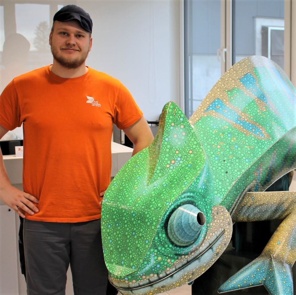 junger Mann, Mütze und orangenes T-Shirt mit Arbeitshose. Arme in die Hüfte, lächelt in die Kamera. Recht von ihm steht ein grünes Chamäleon aus Wellpappe.
