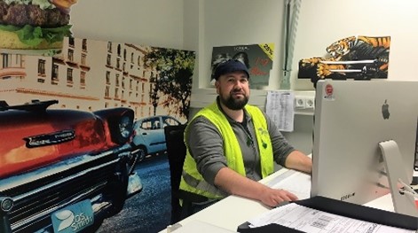 Mann mit Bart, Mütze, Gehörschutz und Sicherheitsweste am Schreibtisch. Er schaut in den PC.